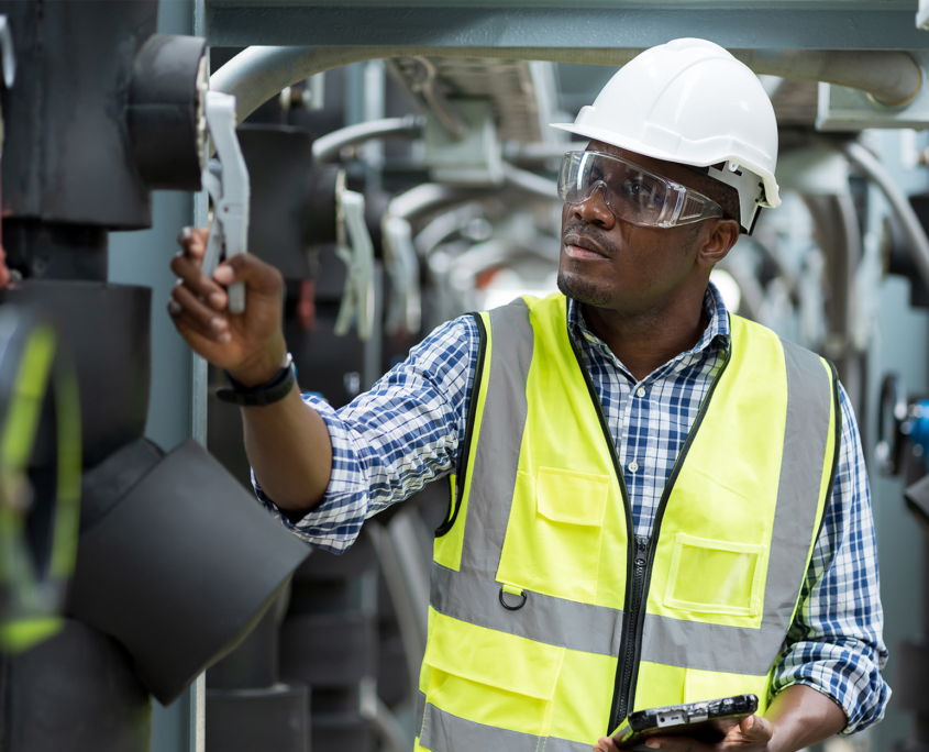 Front view of engineer doing maintenance work at industrial site wearing yellow vest and protective headwear and goggles